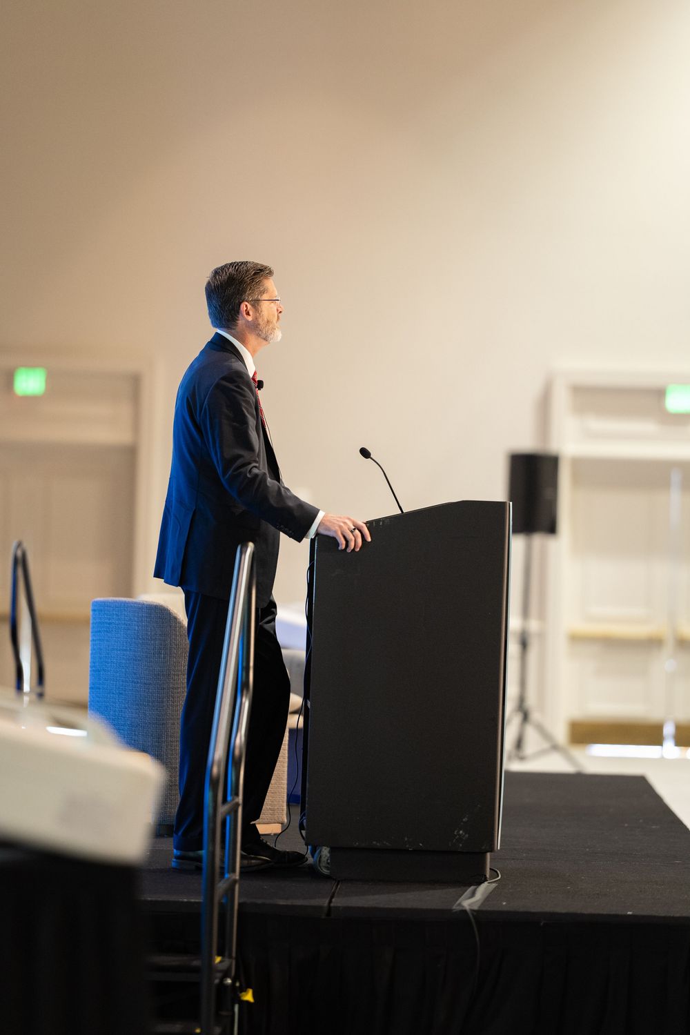A man in a dark suit speaks at a podium with a microphone on a stage in a conference room.