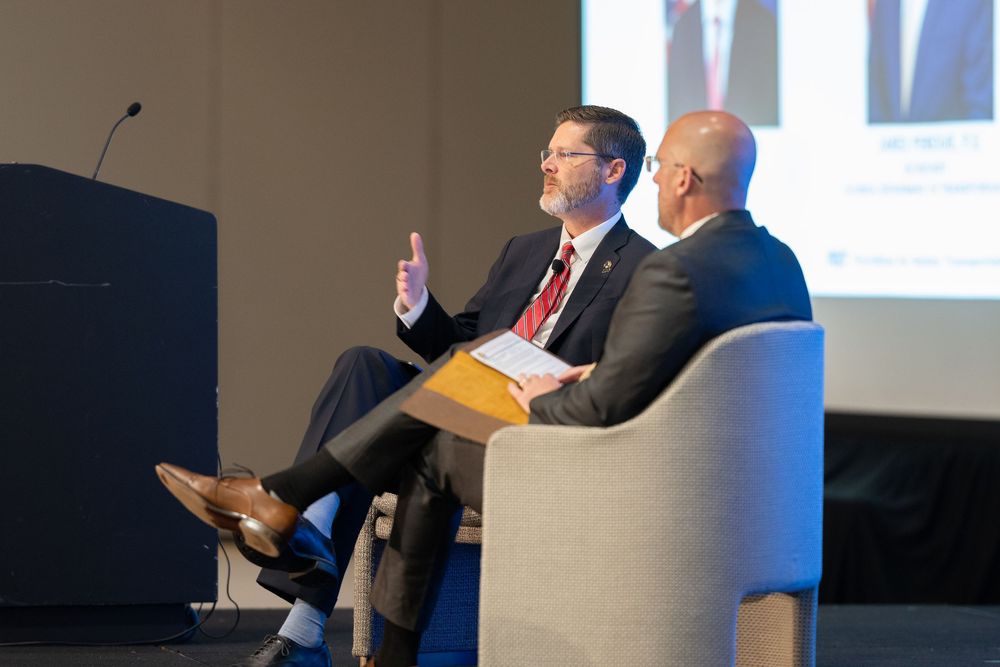 Two business professionals in suits engage in a panel discussion on stage at a corporate conference event.