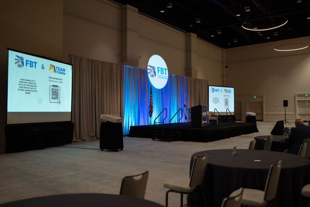 Conference hall with FBT and NAC branded stage, blue lighting, projection screens, and round tables set for an event.