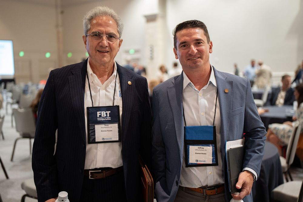 Two professional men wearing conference badges smile together at a formal networking event in a banquet hall.