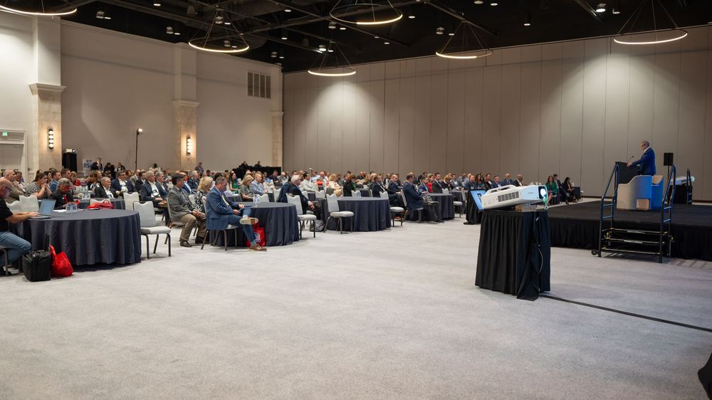 Large conference room filled with attendees listening to a speaker presenting on stage at a professional business event.