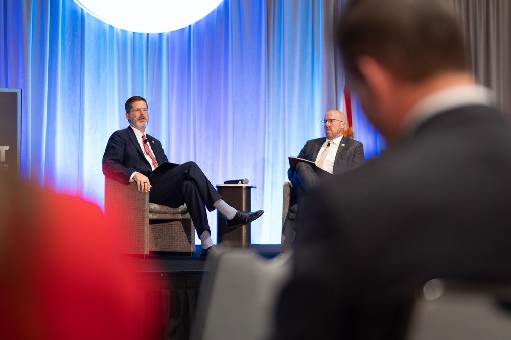 Two businessmen on stage at a corporate conference, one seated in a chair, the other holding notes at a podium.