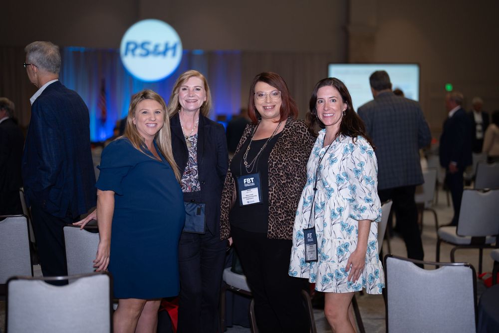 Four women smiling together at an RS&H corporate event in a conference hall with blue branded backdrop.
