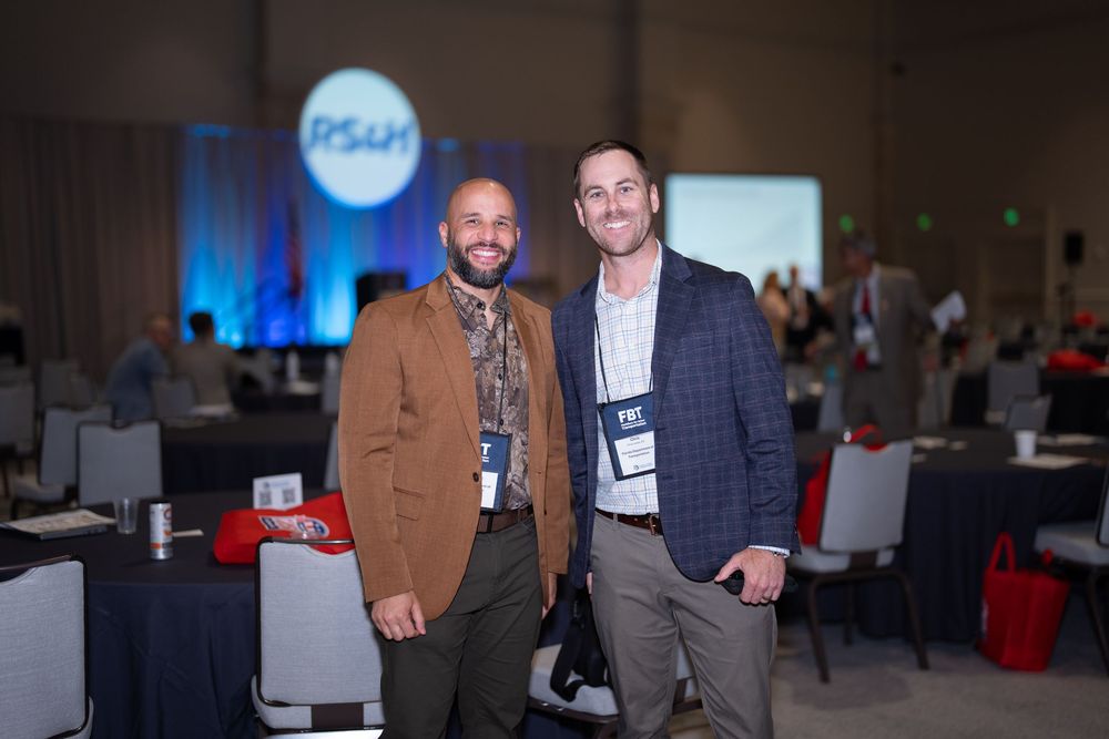 Two men in business casual attire smile at an RS&H conference event in a large banquet hall.