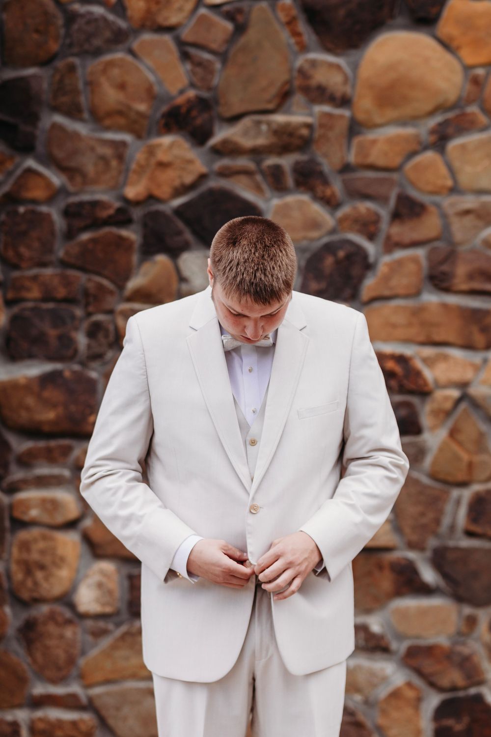 Groom in white suit buttoning jacket against rustic stone wall backdrop.