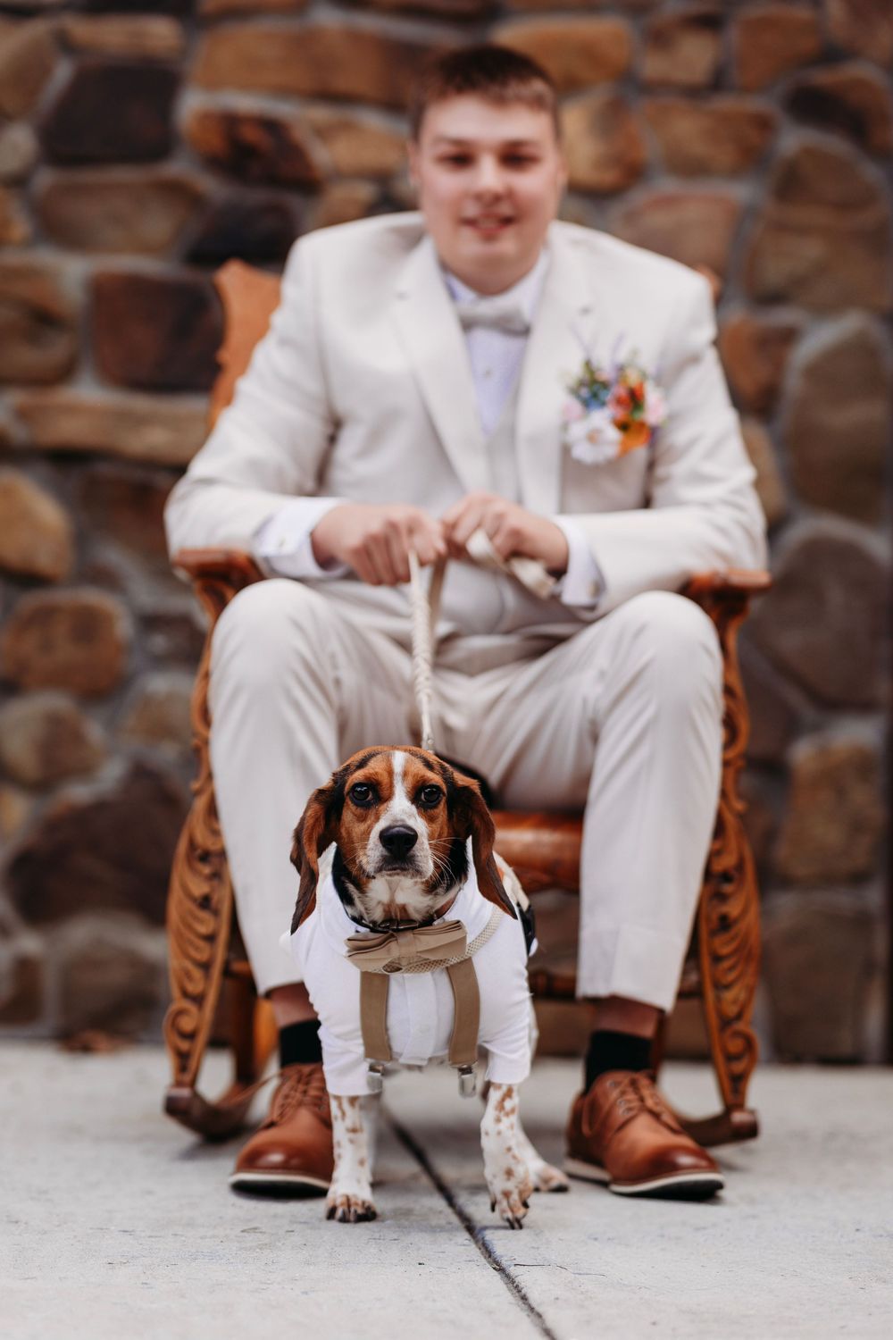 A smiling groom in a white suit sits in a wooden chair holding a leash attached to a dressed beagle dog.