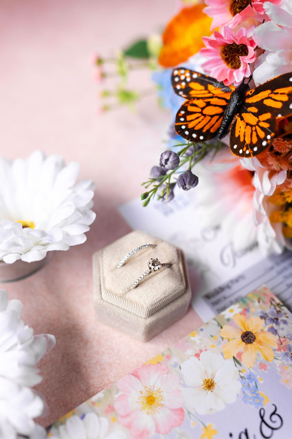 Diamond engagement ring in velvet box surrounded by colorful flowers and a monarch butterfly on pink background.