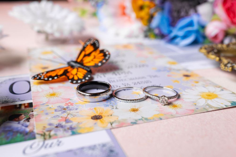 Wedding rings and diamond engagement ring displayed on floral invitation with monarch butterfly decoration.