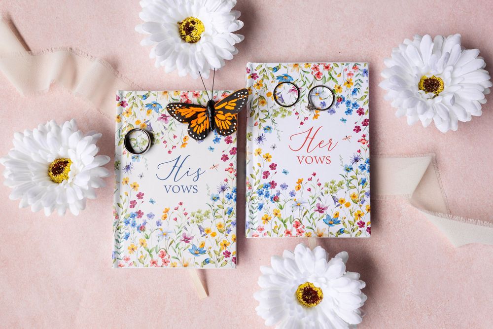 Floral his and her vows books with wedding rings, cufflinks, and a monarch butterfly on a pink background.