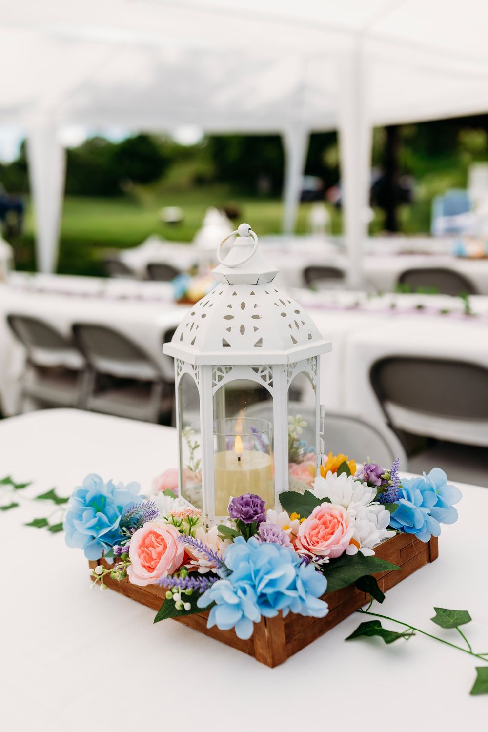 White lantern centerpiece with lit candle surrounded by colorful flowers on wooden tray at outdoor wedding reception.