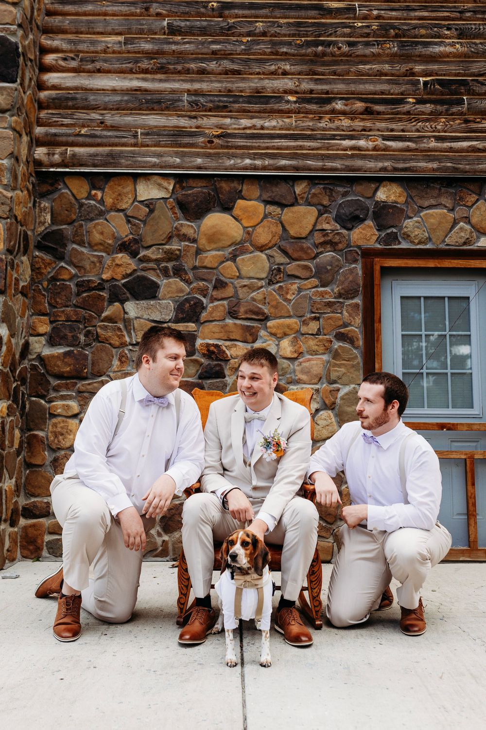 Groom and groomsmen in white shirts pose with a beagle dog outside a rustic stone and log cabin venue.