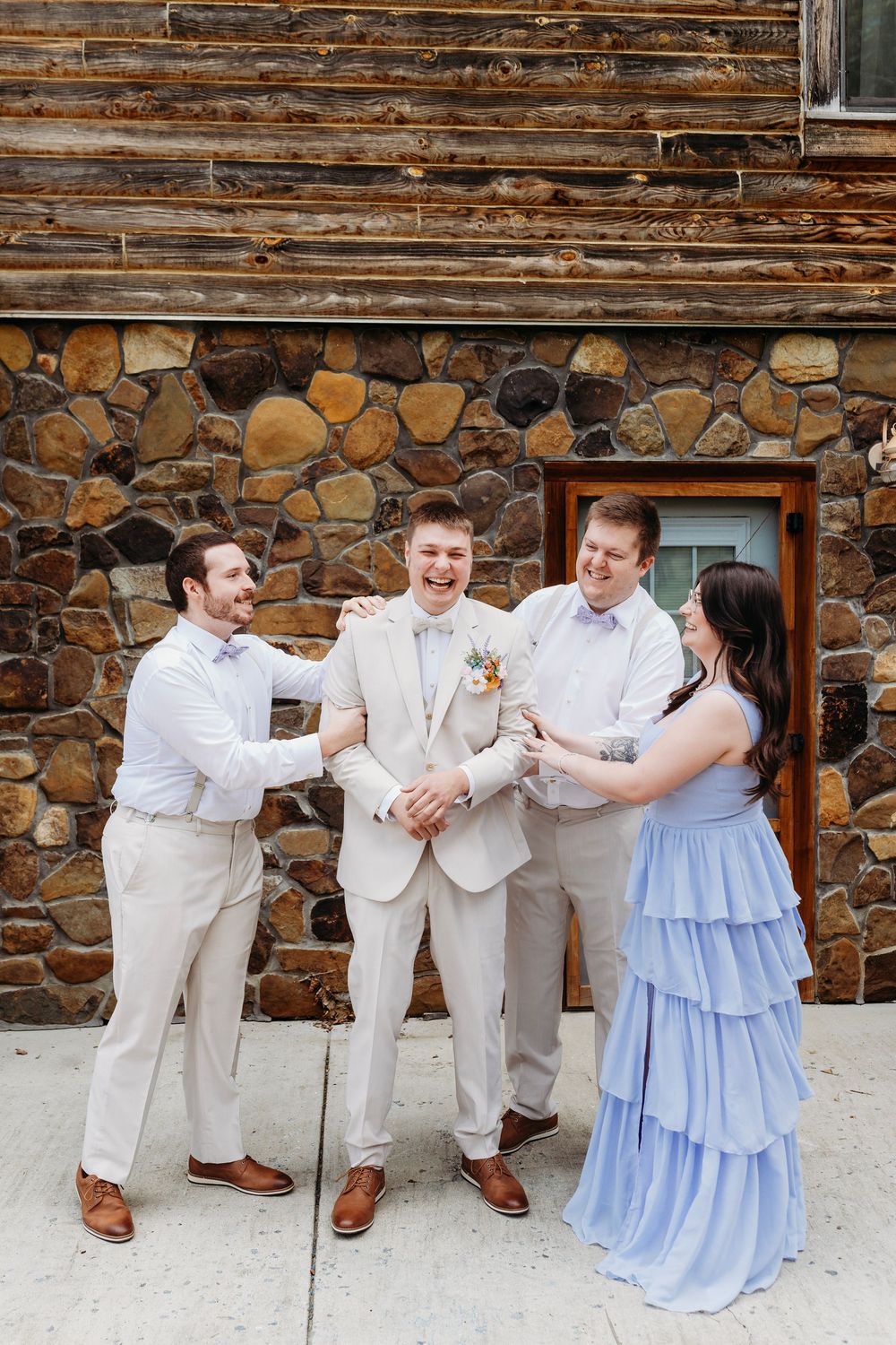 Groom in beige suit laughs with groomsmen in white shirts and bridesmaid in blue gown outside stone building.
