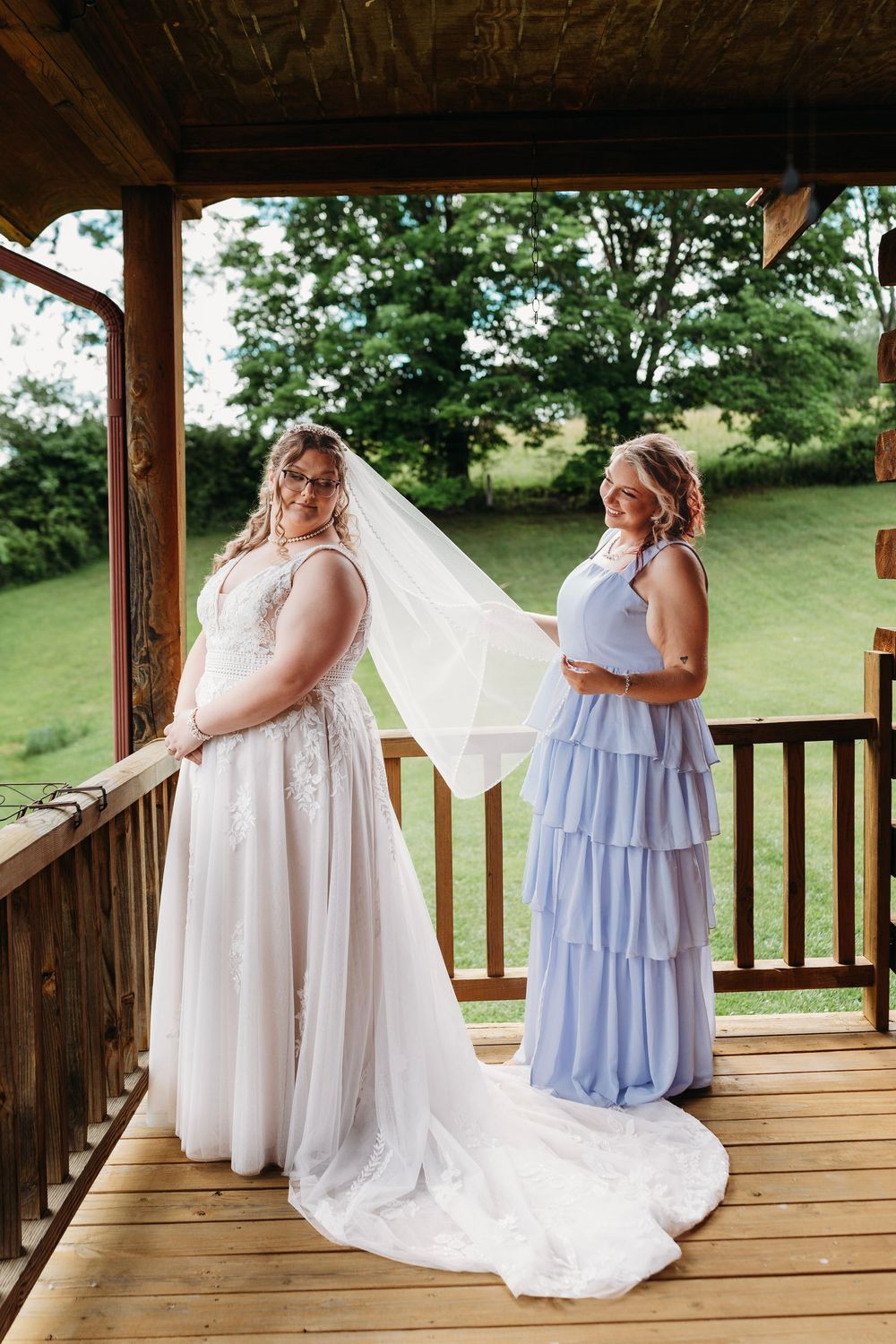 Bride in white lace gown with veil and bridesmaid in blue dress on wooden porch at outdoor wedding venue.