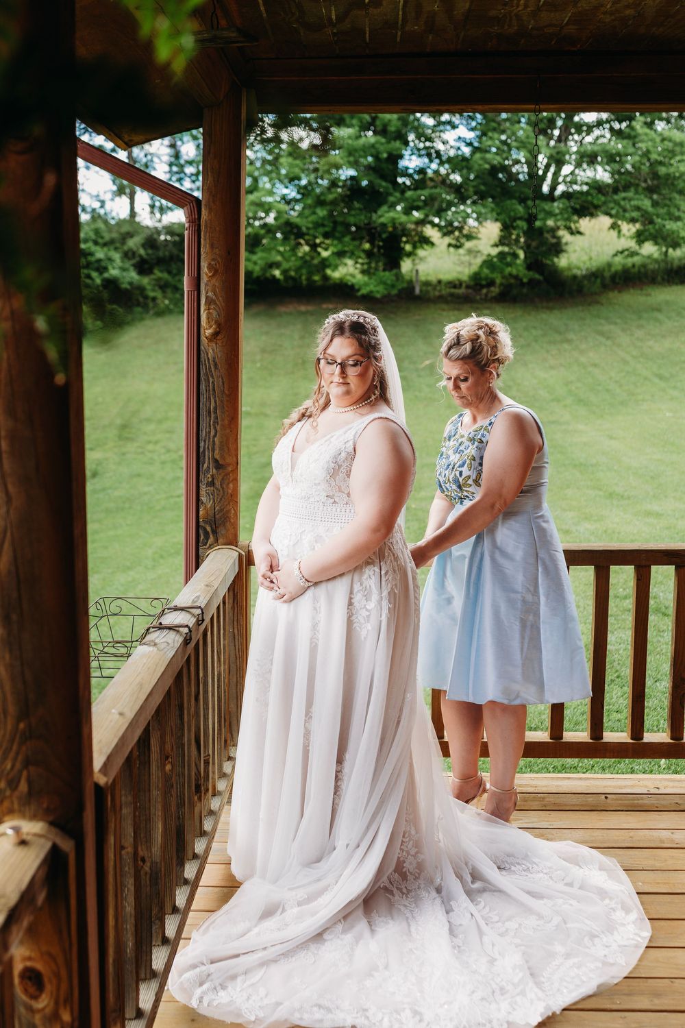 Bride in white lace gown stands on wooden deck while bridesmaid in blue dress buttons up the back of her dress.