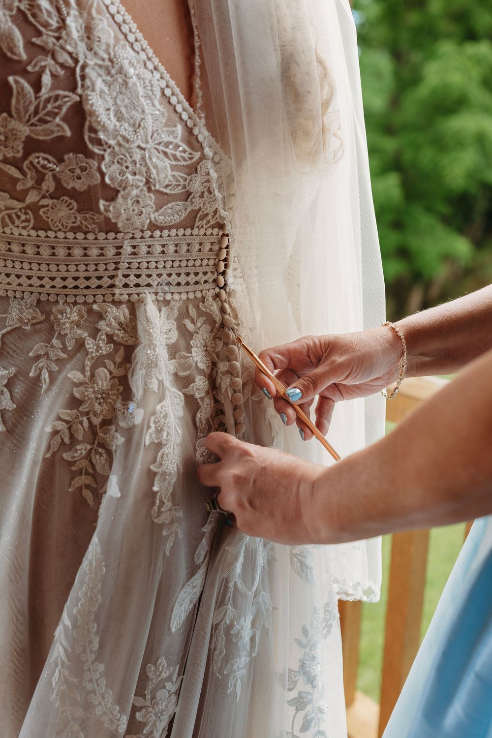 Hands fastening buttons on a bride's elegant white lace wedding dress with intricate floral embroidery.