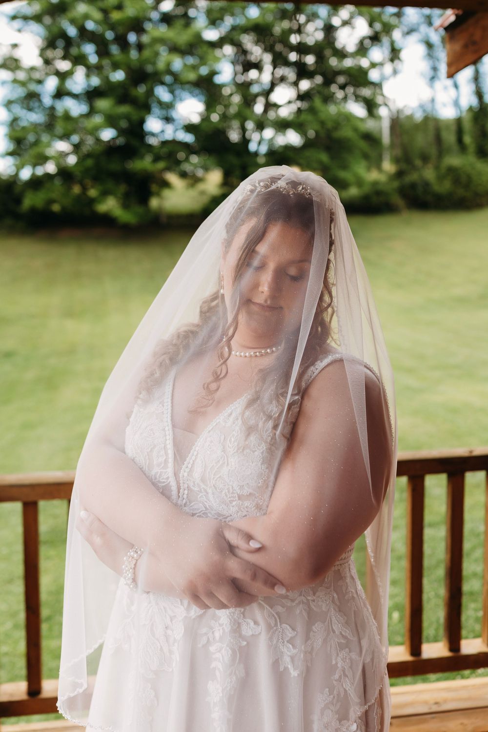 Bride in lace wedding dress and veil looking down with arms crossed outdoors at garden wedding venue.