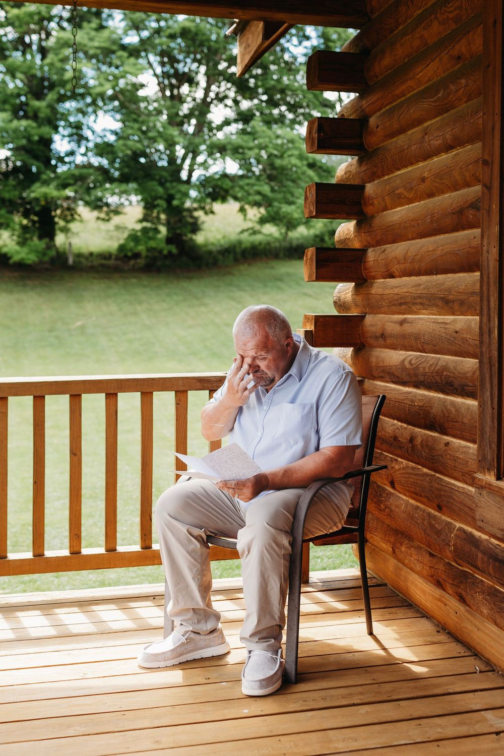 Elderly man sitting on wooden cabin porch reading a letter with hand on face, surrounded by lush green trees.