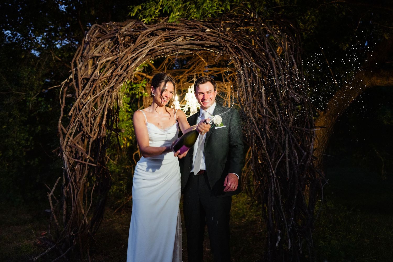 Bride and groom smiling under a rustic twig arch outdoors at their wedding reception.