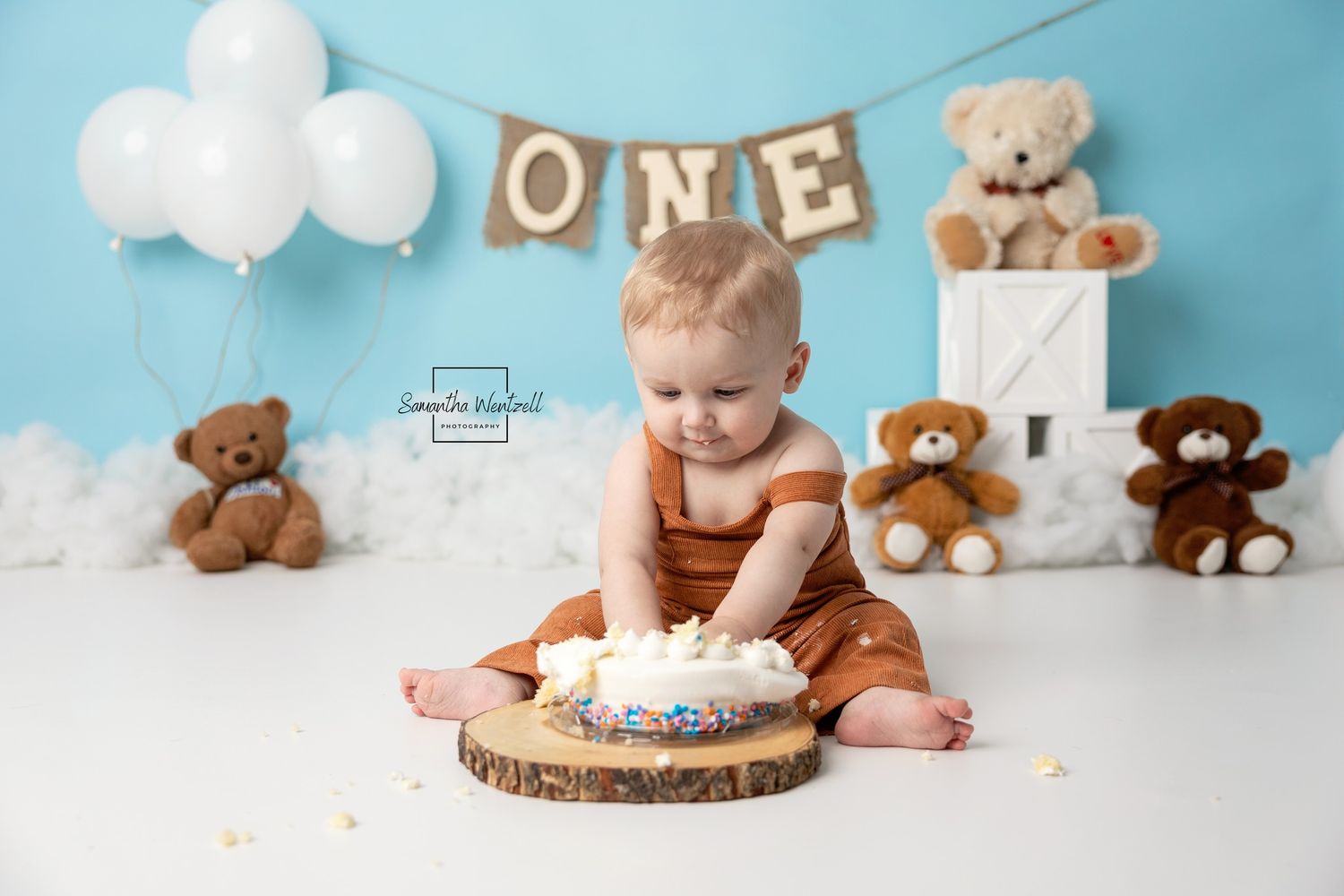 Baby in rust romper smashes first birthday cake surrounded by teddy bears and balloons under ONE banner.