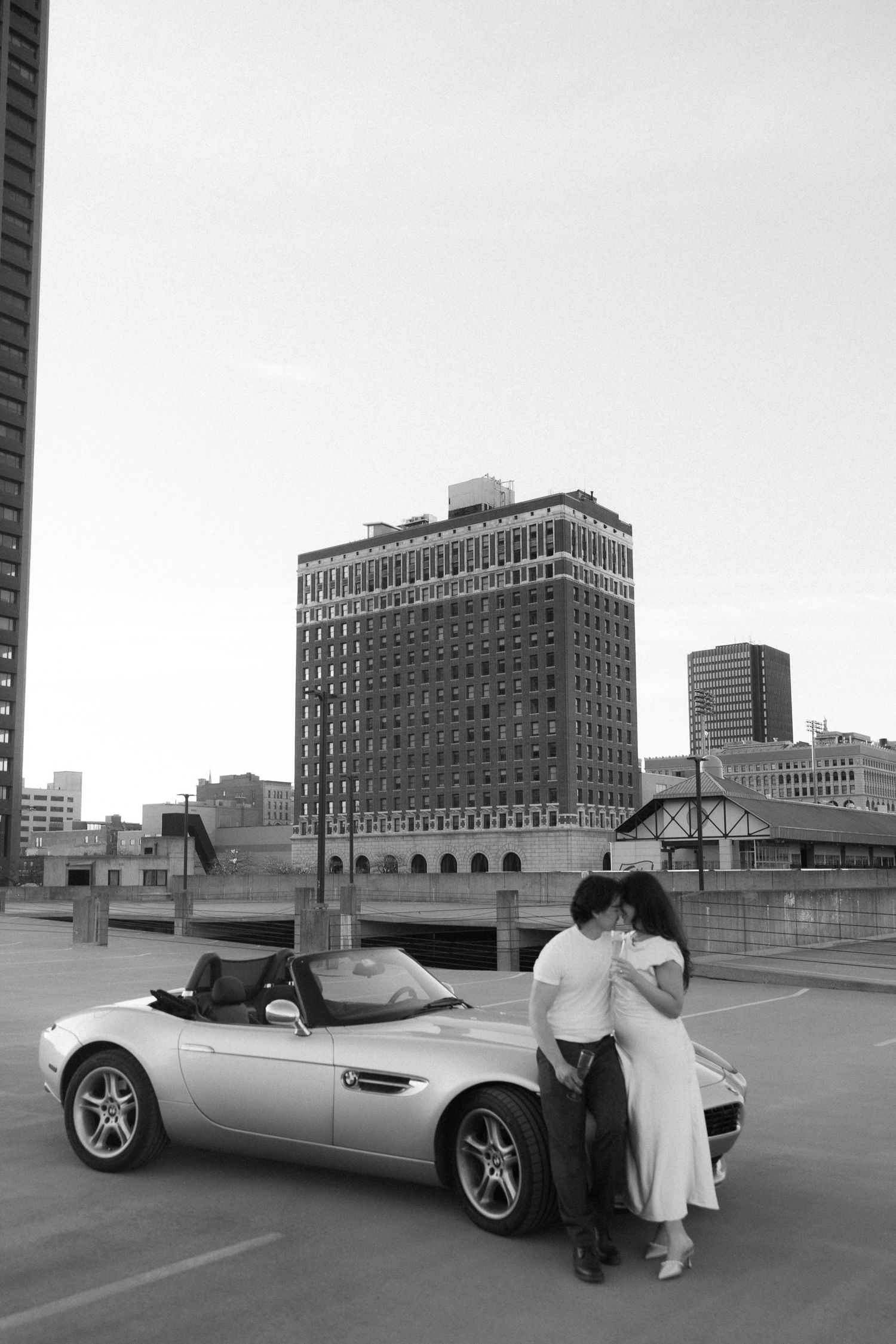 Black and white photo of a couple embracing beside a silver convertible sports car in an urban city setting.