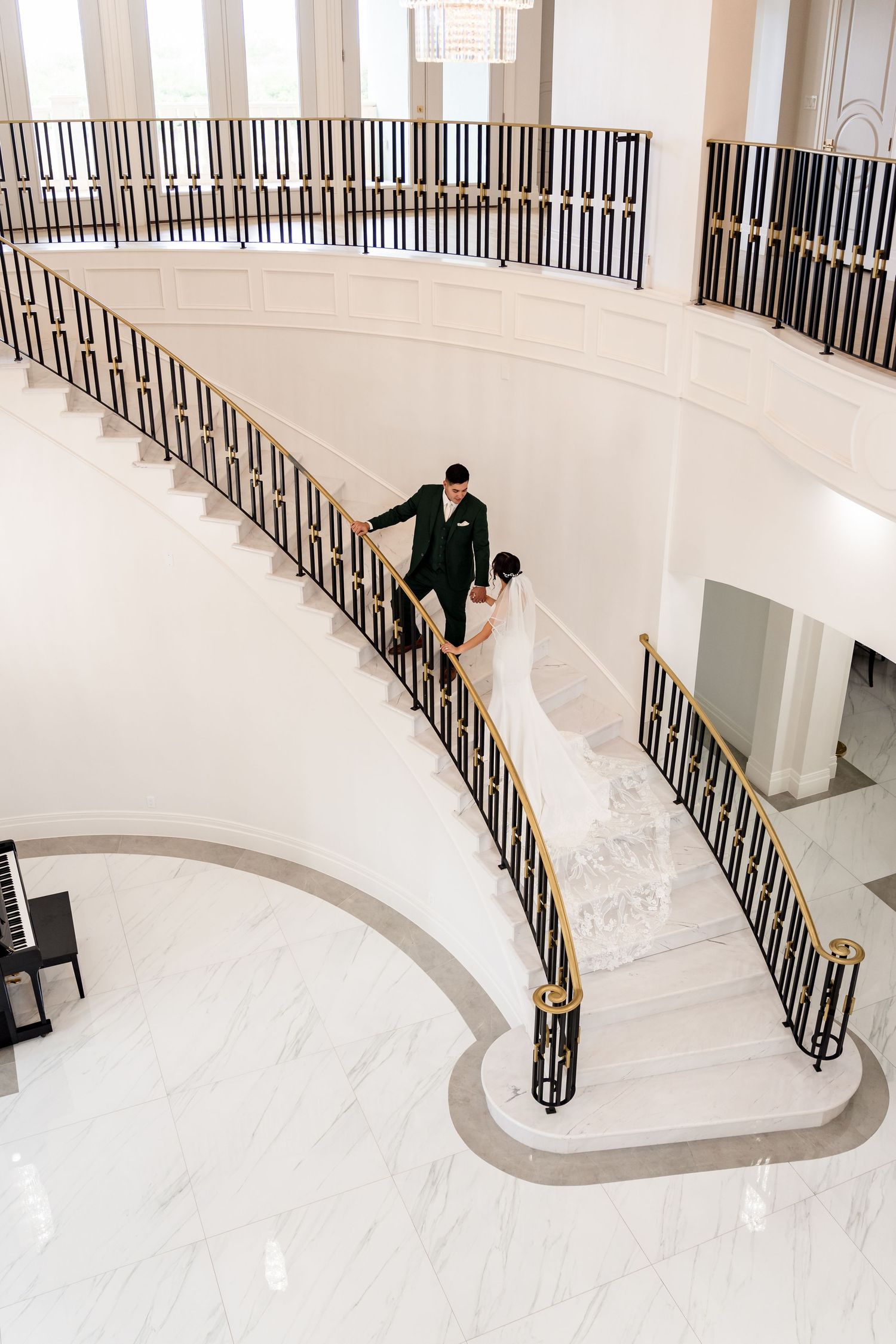 Bride and groom descend a grand curved marble staircase with gold railings in an elegant white wedding venue. RGV Photoshoot.
