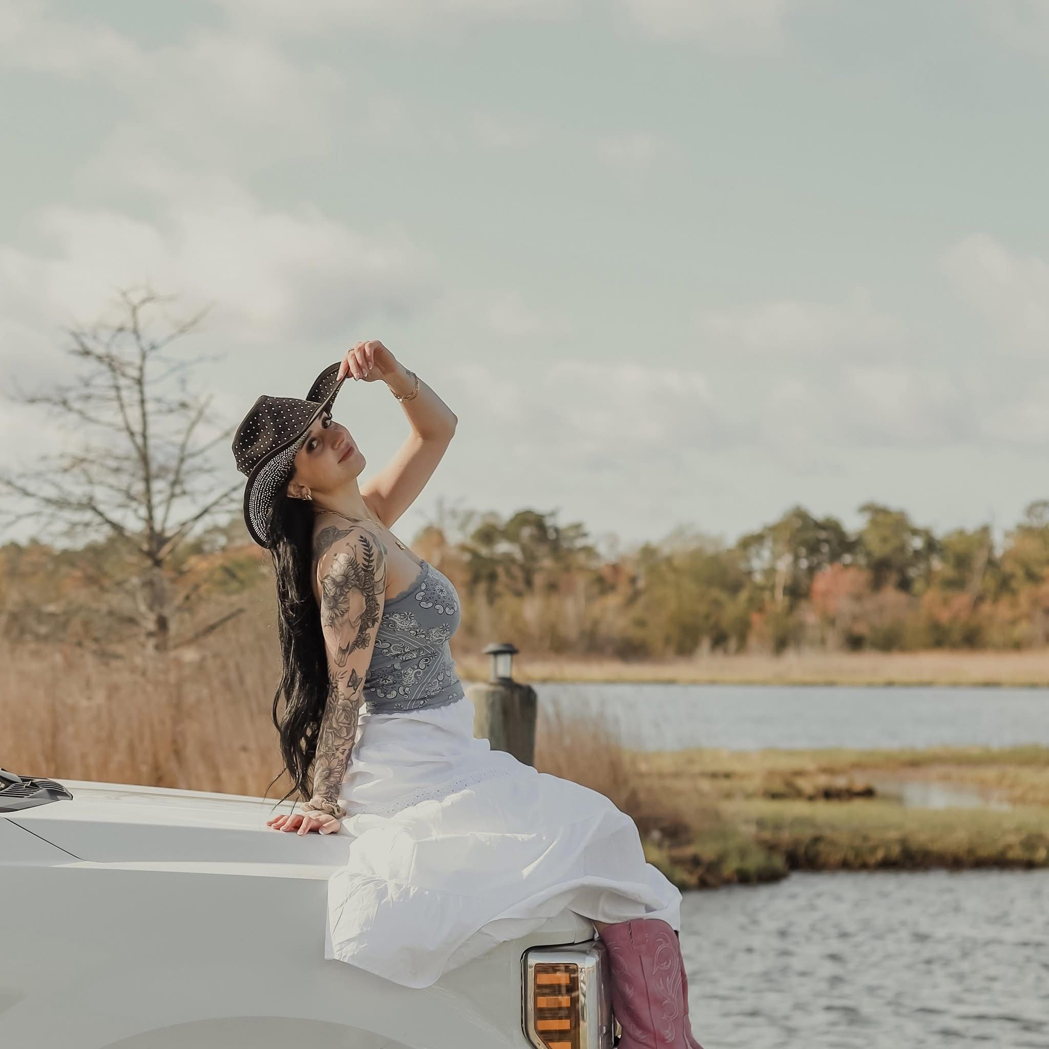 Tattooed woman in cowboy hat and white pants sitting on white vehicle near a serene marsh landscape.