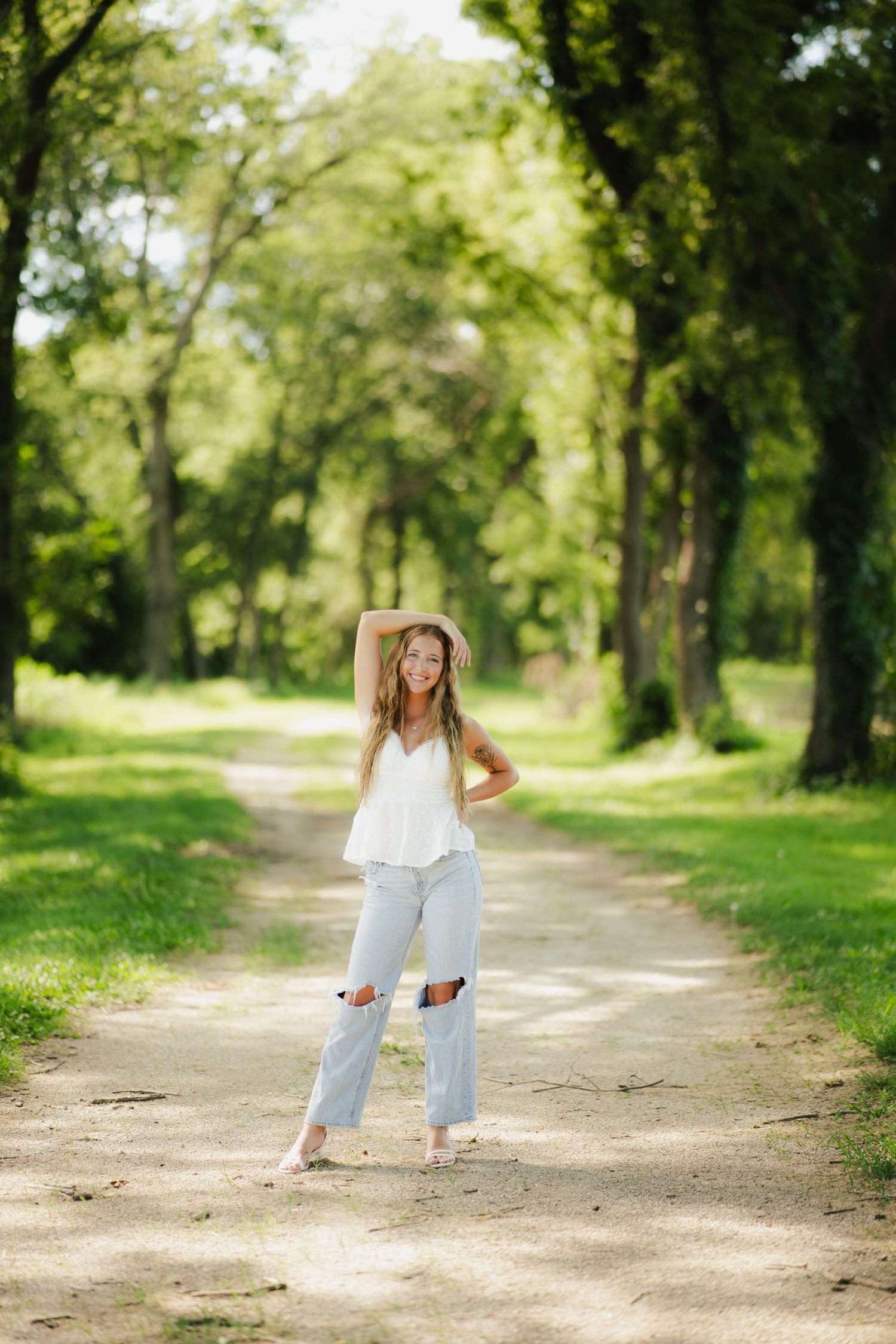 Young woman in white top, ripped jeans, and straw hat posing on a tree-lined dirt path in a sunny green park.