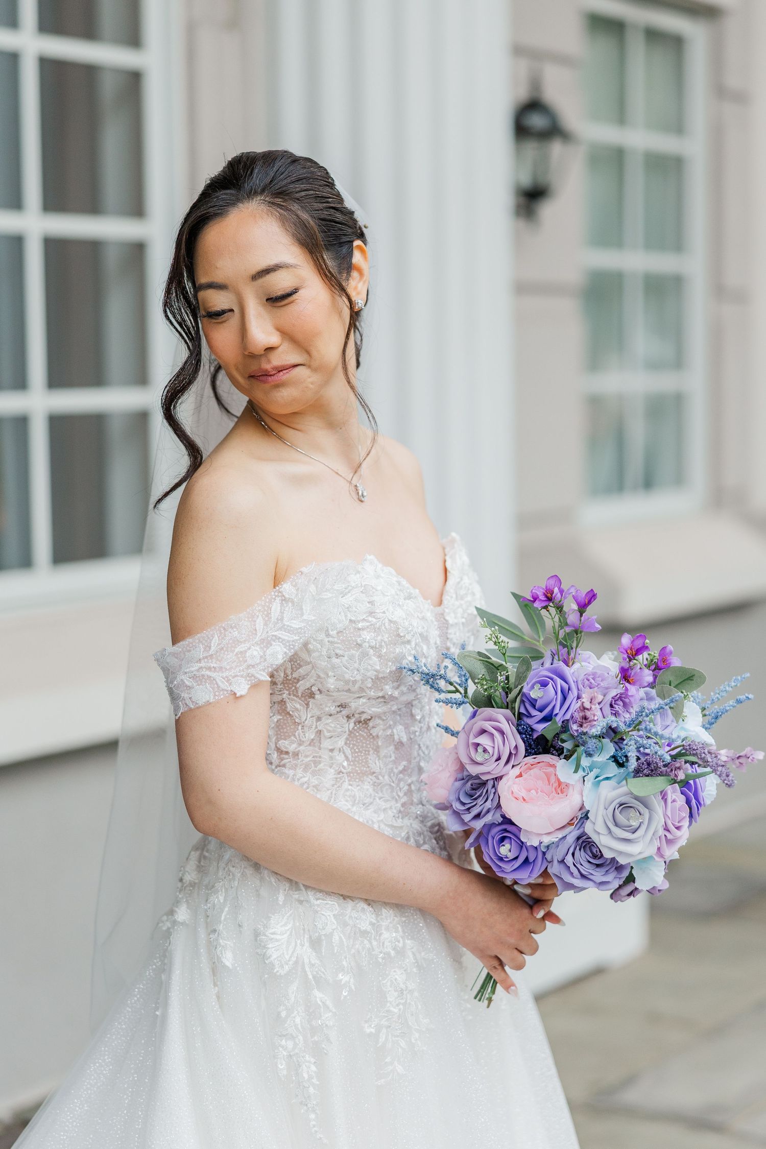 Bride in off-shoulder lace gown holding purple and pink floral bouquet, posing elegantly outdoors.