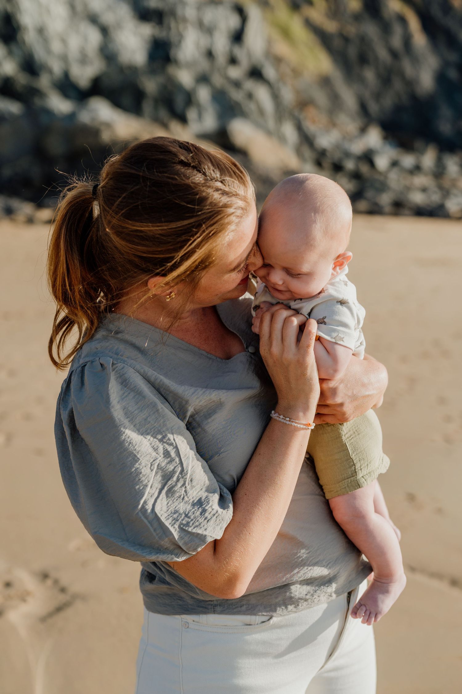 A mother tenderly holds and nuzzles her newborn baby on a sandy beach with rocky cliffs in the background.