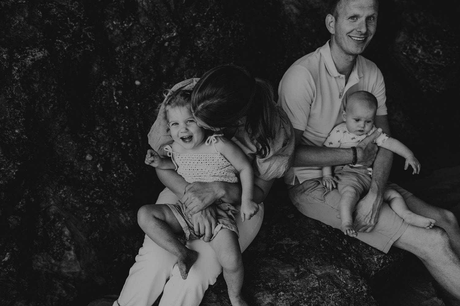 Black and white family portrait of parents with two young children sitting on rocks, sharing a joyful moment outdoors.