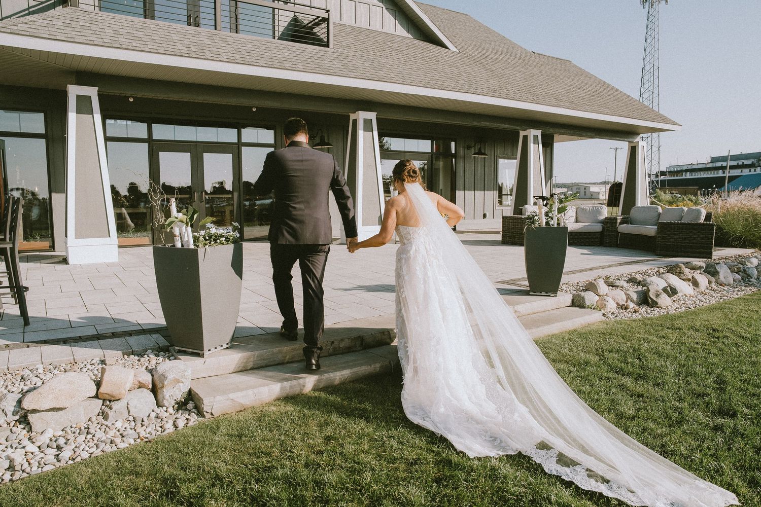 Bride and groom walking hand in hand toward elegant wedding venue with manicured grounds and modern architecture.