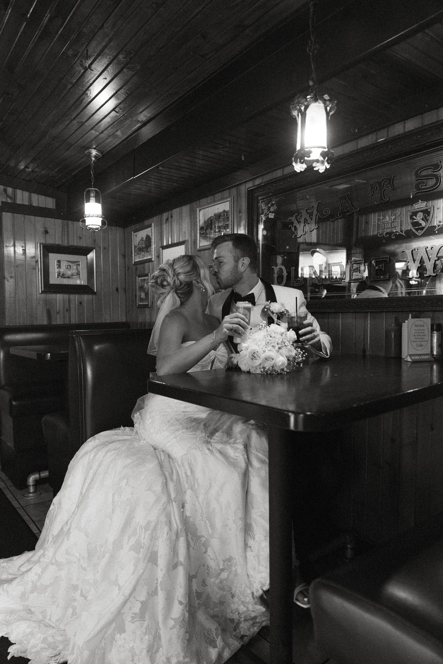 Black and white photo of a bride and groom sharing a romantic moment at a wooden bar, bride holding a bouquet.