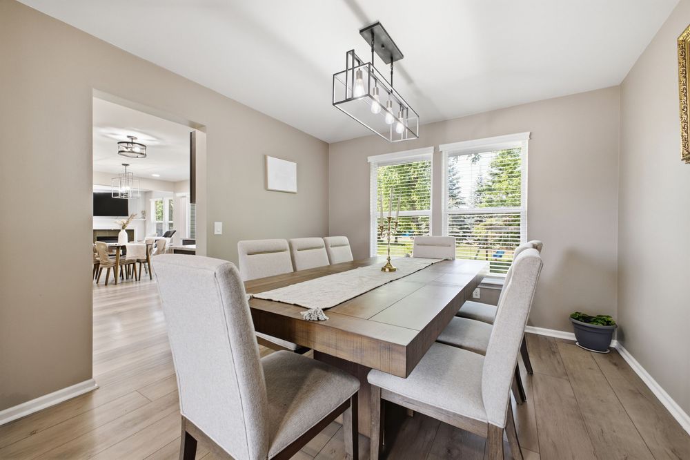 Bright dining room with wooden table, white upholstered chairs, modern chandelier, and large windows overlooking greenery.