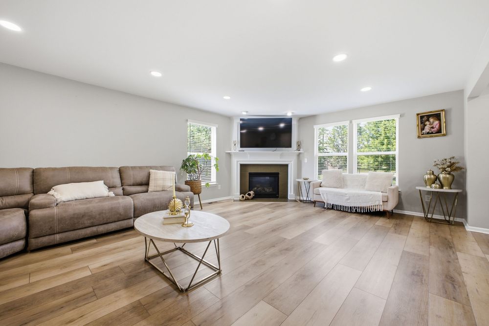 Bright living room with hardwood floors, fireplace, brown leather sofa, and large windows providing natural light.