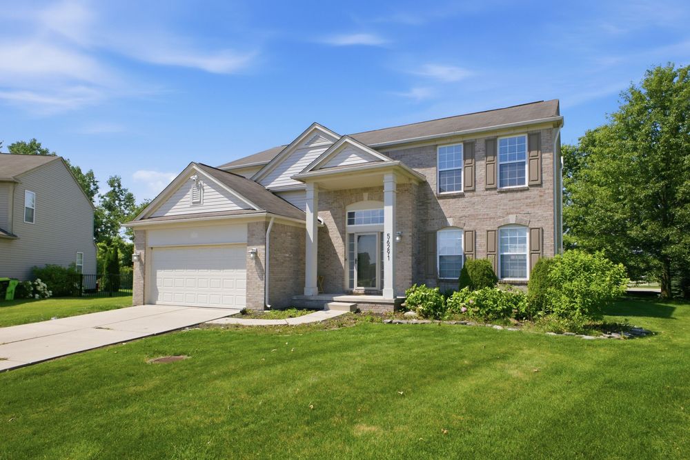 Two-story brick colonial home with attached garage, covered entry, and lush green lawn surrounded by mature trees.