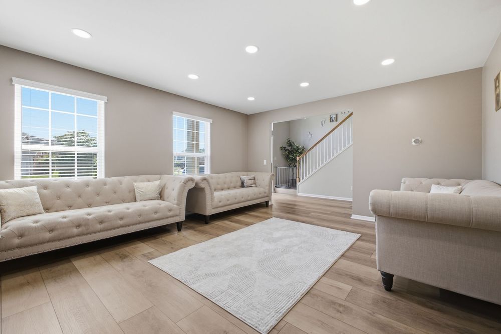 Bright living room with beige tufted sofas, light hardwood floors, white rug, recessed lighting, and staircase.