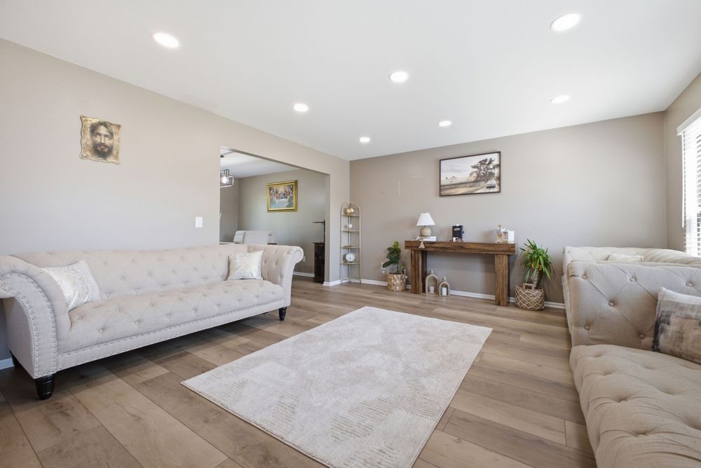 Bright living room with beige tufted sofas, hardwood floors, a white area rug, and a decorative fireplace mantel.