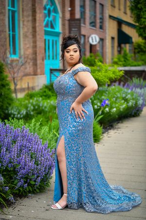 Elegant young woman in blue lace formal gown standing beside purple flowers outdoors.