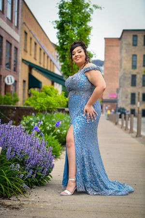 Young woman in blue sequin prom gown posing near purple flowers on urban walkway.