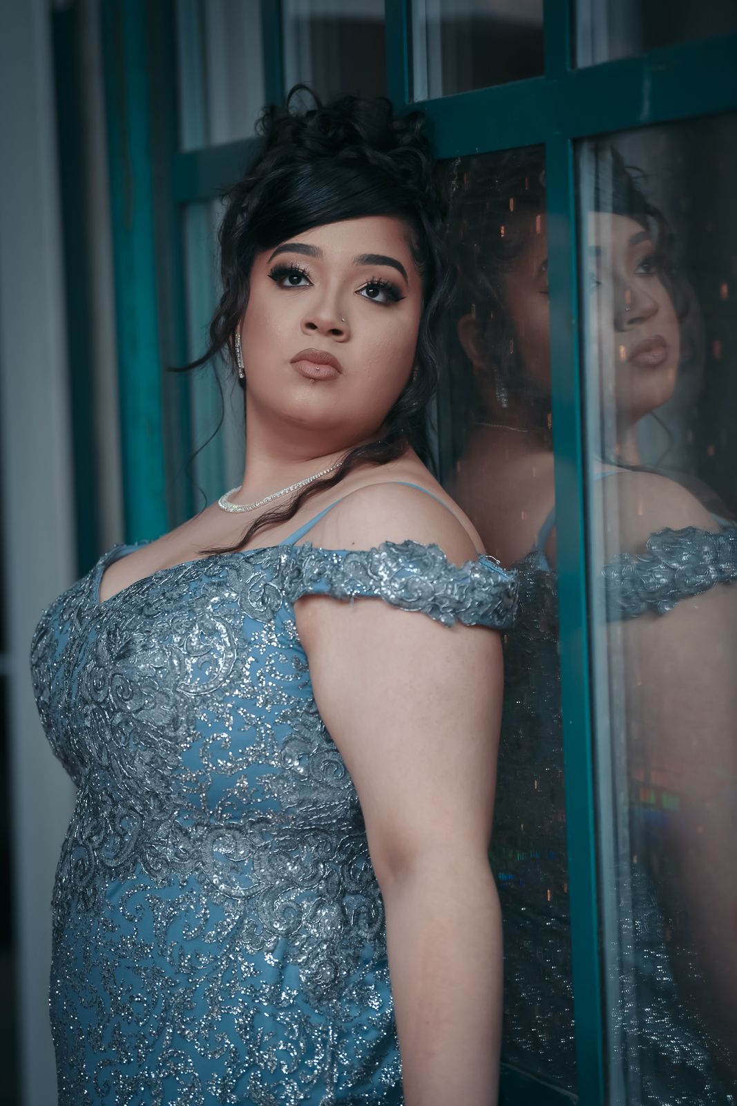 Young woman in blue sequin off-shoulder gown posing against teal window with reflection.