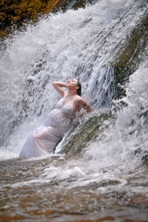 Pregnant woman in white dress stands in rushing waterfall with head tilted back in serene moment.