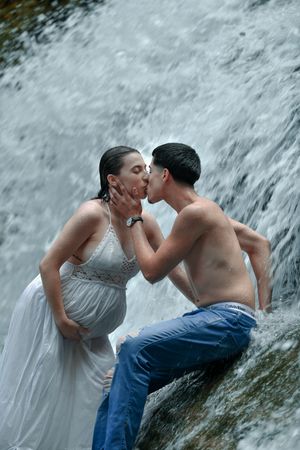 Couple shares romantic kiss beside cascading waterfall, woman in white dress, man shirtless in jeans.