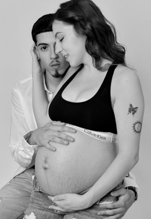 Black and white maternity photo of couple embracing pregnant woman's bare belly in studio setting.