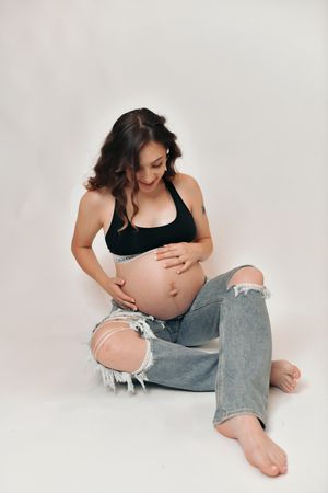 Smiling pregnant woman in black sports bra and ripped jeans sits barefoot cradling her baby bump.