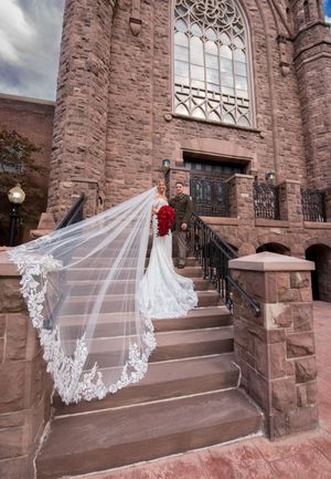 Bride in lace gown with cathedral veil and groom on stone steps of historic church wedding venue.