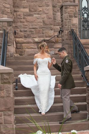 Military groom in green uniform escorts bride in off-shoulder lace dress up stone church steps.