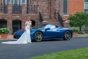 Elegant bride in white mermaid gown posing beside a blue Ferrari outside a brick mansion.