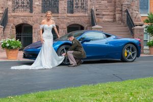 Bride in white gown stands with groom crouching by blue Ferrari sports car at brick estate.