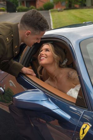 Joyful bride in lace gown laughing with military groom leaning into blue Ferrari car window.