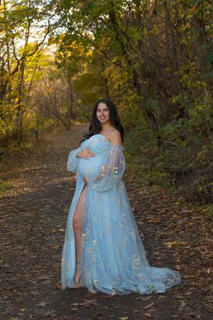 Smiling pregnant woman in floral light blue tulle gown poses on autumn forest path at golden hour.